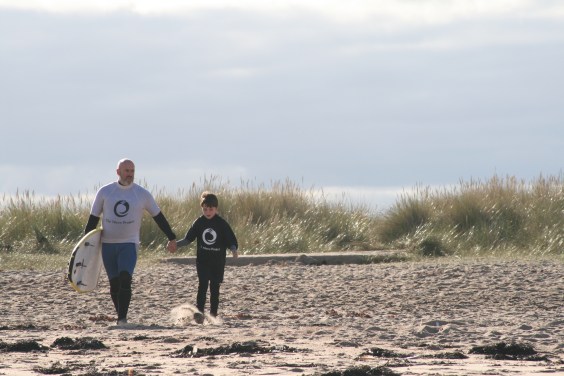 A man carrying a surf board holding the hand of a young boy and walking across a sandy beach. 