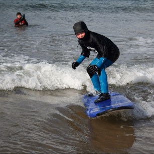 A young person surfing in the sea with a support worker behind him