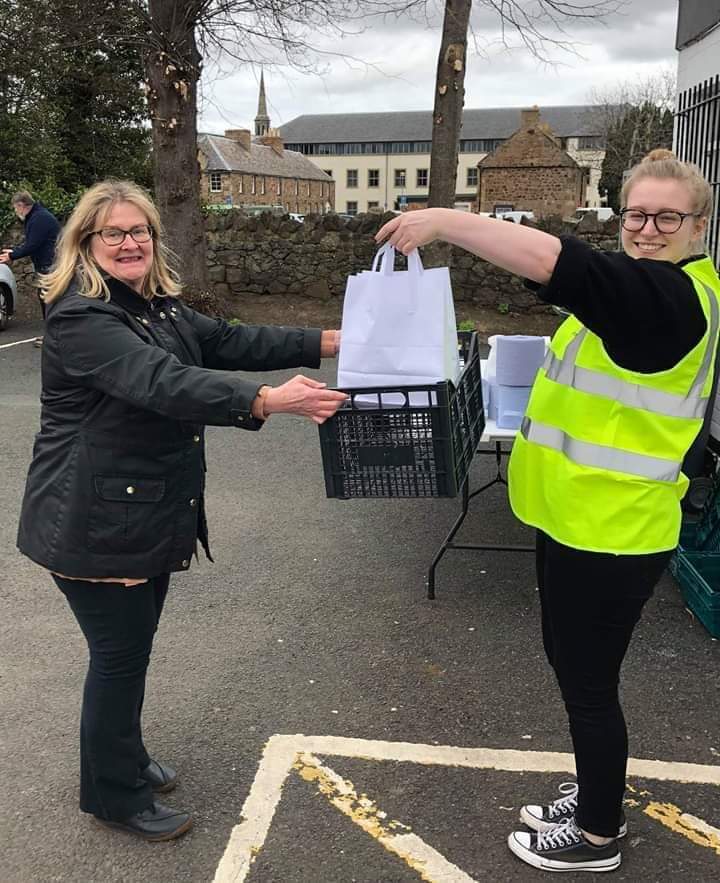 One volunteer putting a paper delivery bag into a plastic crate that the other volunteer is holding. Both smiling.