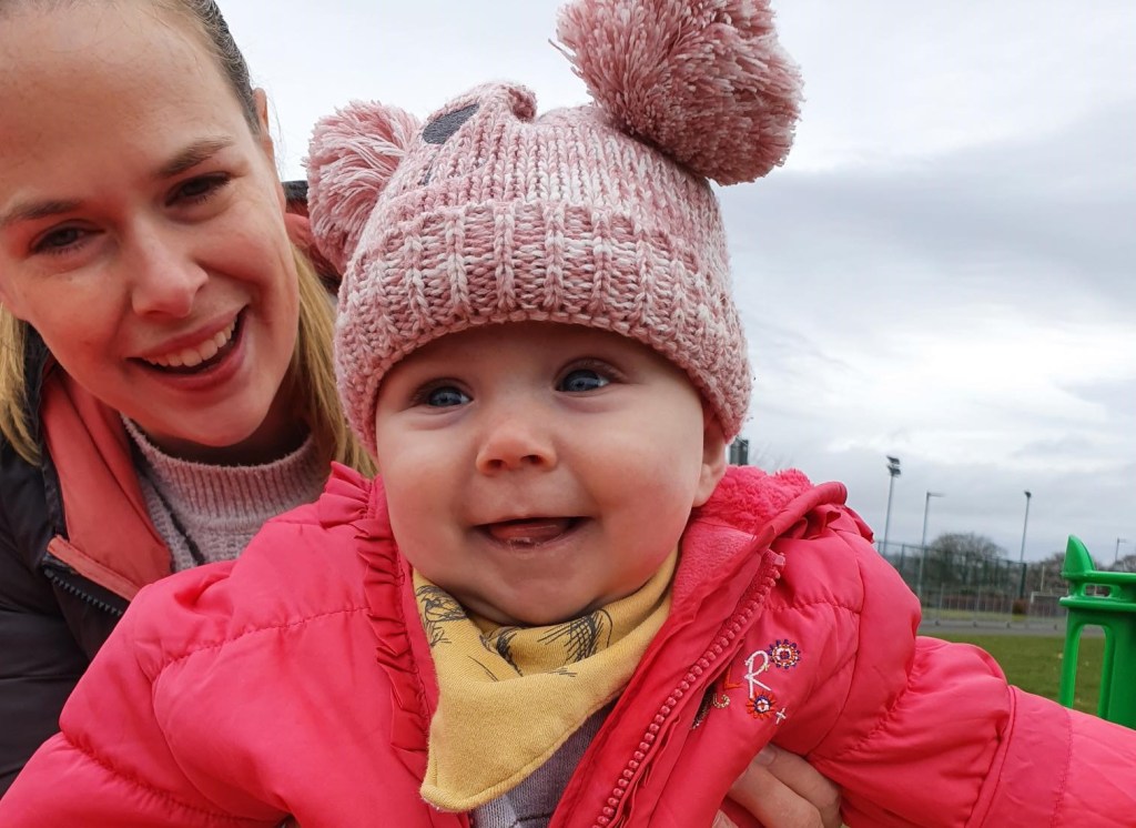 Smiling Baby Eryn wearing a red coat and pink pom-pom hat, while mum Jane holds her up from behind.