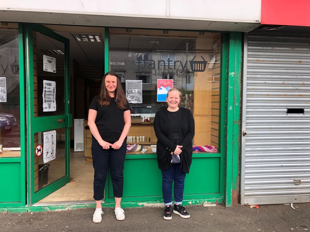 Two volunteers stand outside the community pantry smiling.