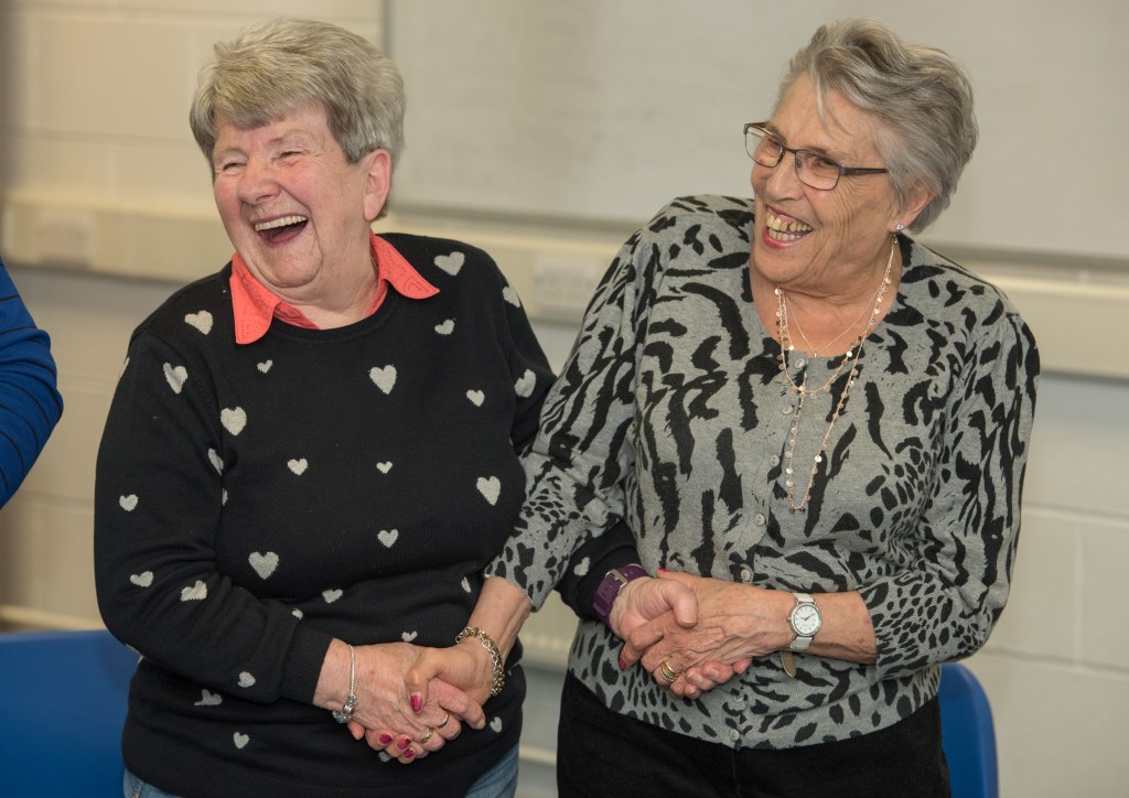 Two members of The Warblers smiling and laughing as they hold hands as they sing and have a laugh with the group. Another image taken before the pandemic.