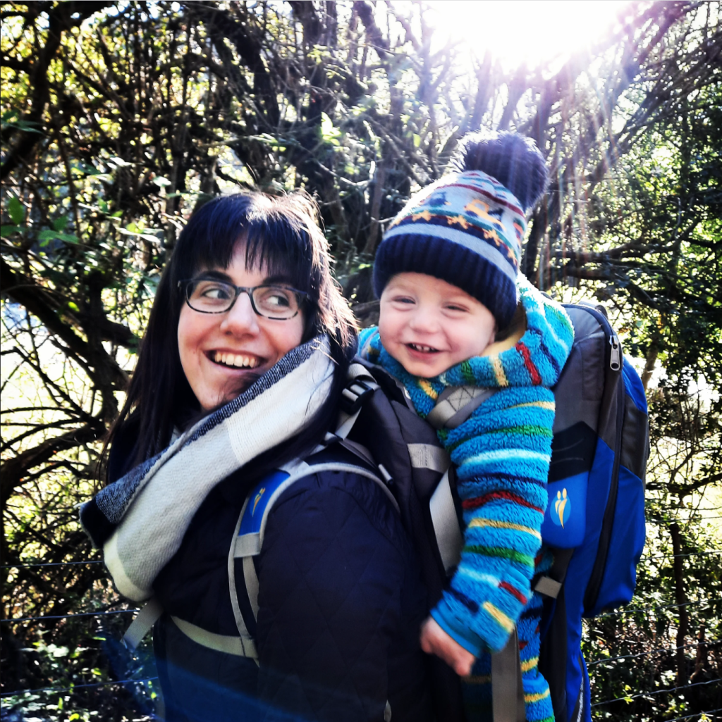 Laura is smiling as she walks in a woodland area, she wears a scarf and a rucksack carrier with her son Reuben in it. He is smiling, wrapped up in a cosy onesie and hat. 