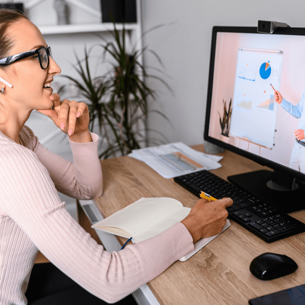 someone takes notes at a desk as they listen to an online workshop.