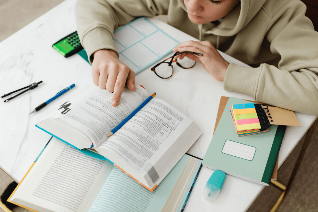 Image from a birds-eye view shows a student reading a textbook. The desk is filled with post it notes, highlighters and notebooks as they study.