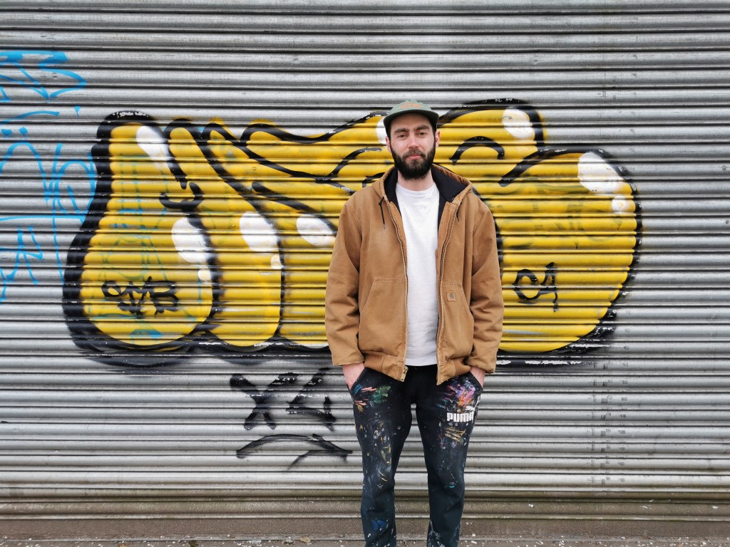 picture of panda, with his hands in his pockets, smiling at the camera. He stands in front of street art on a garage door.