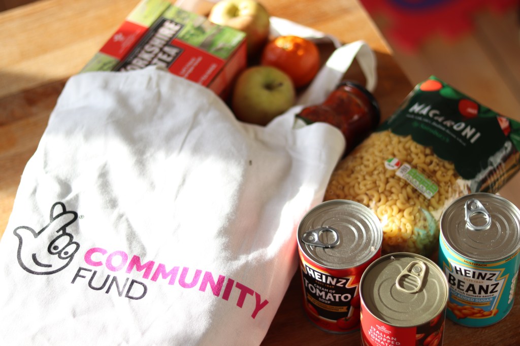 A canvas bag with The National Lottery Community Fund's logo on it is laid out on a table. There are tins of soup, apples and pasta next to it on the table.