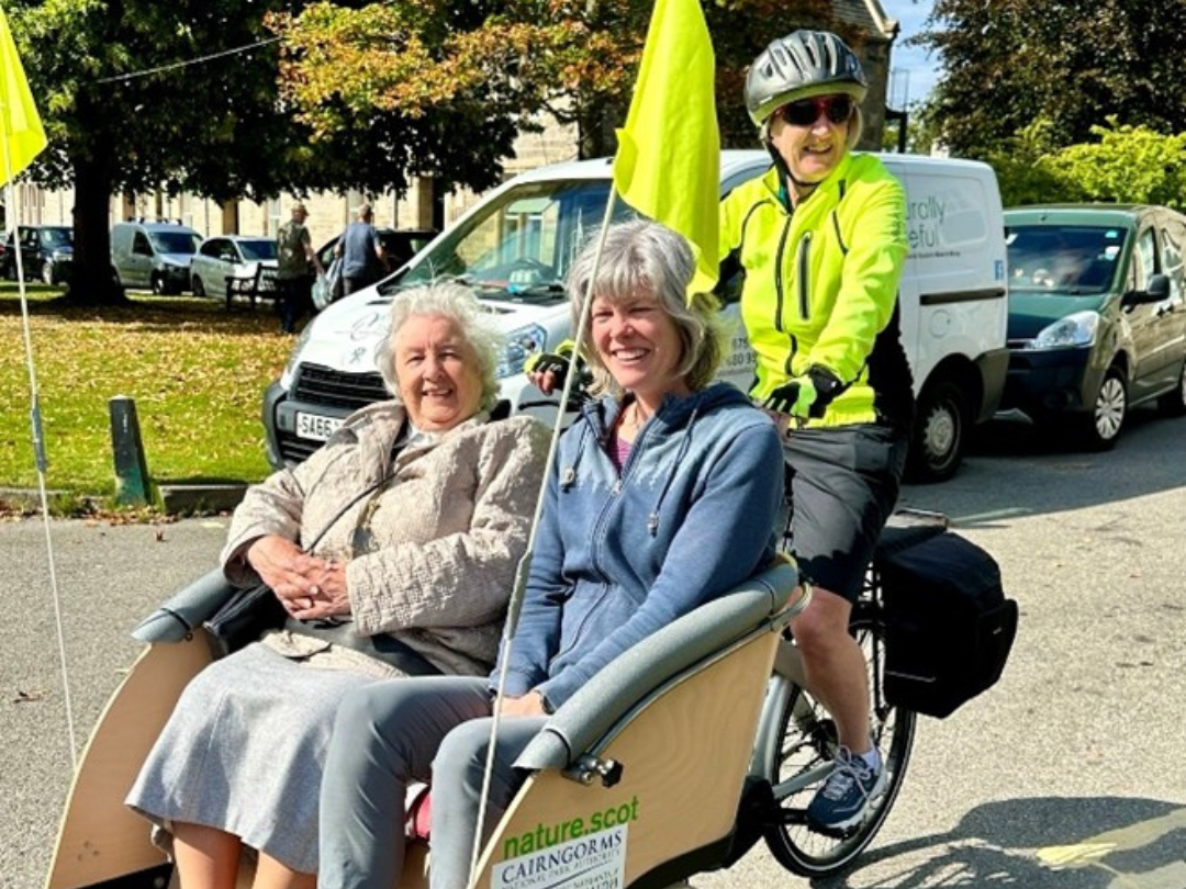 Two people smile as they sit on an accessible bike whilst someone cycles behind them.